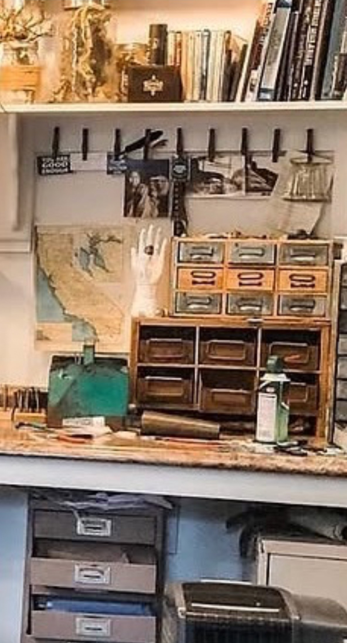 Jenna's actual workbench in the Mount Shasta studio — a small oak drawer cabinet standing on the bench, a white display hand wearing a ring, a map pinned to the wall, photographs clipped along a wire above, grey flat-file drawers below