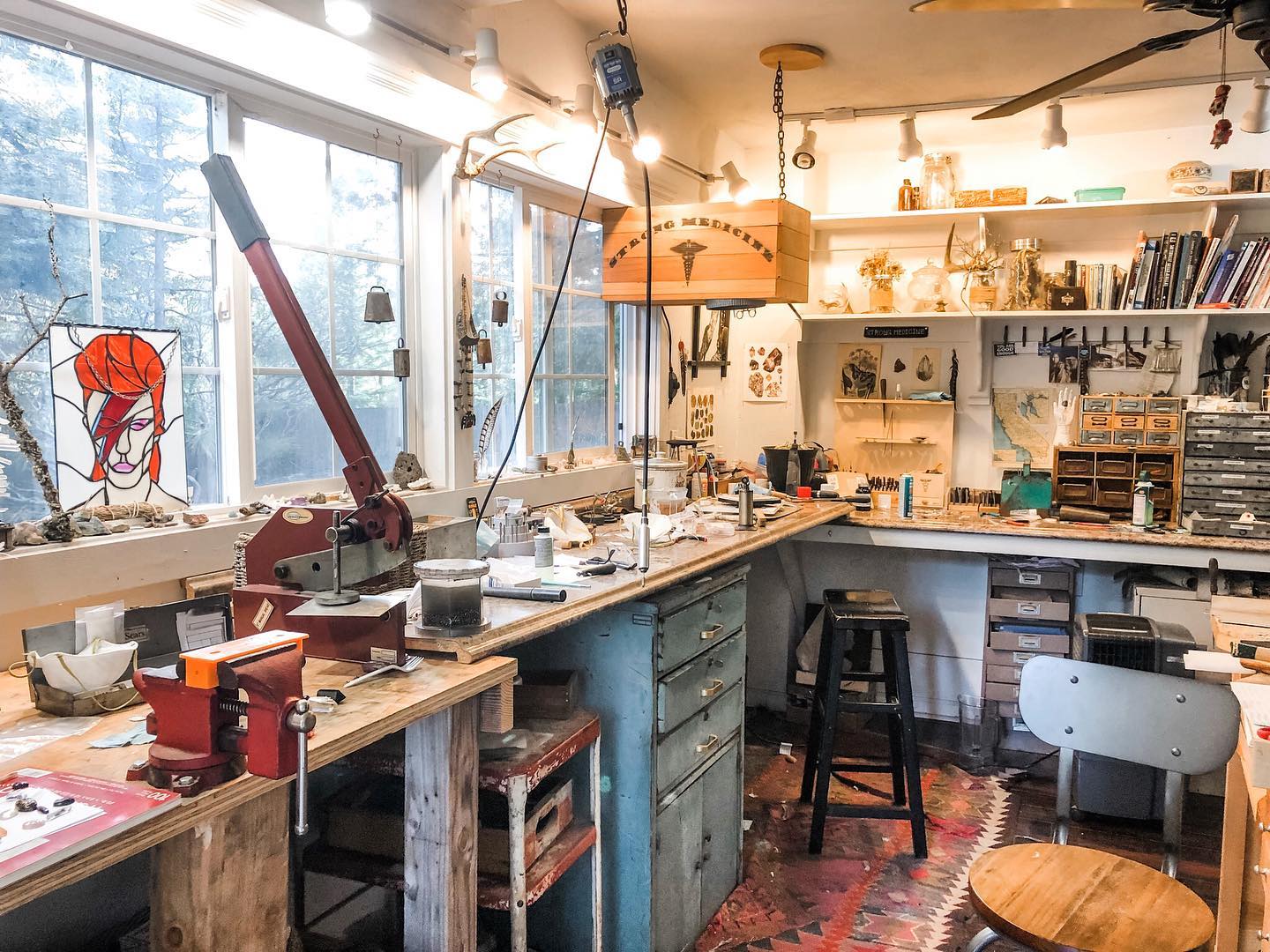 Inside the studio — Jenna's workbench under tall windows, the wooden Strong Medicine sign hanging above it
